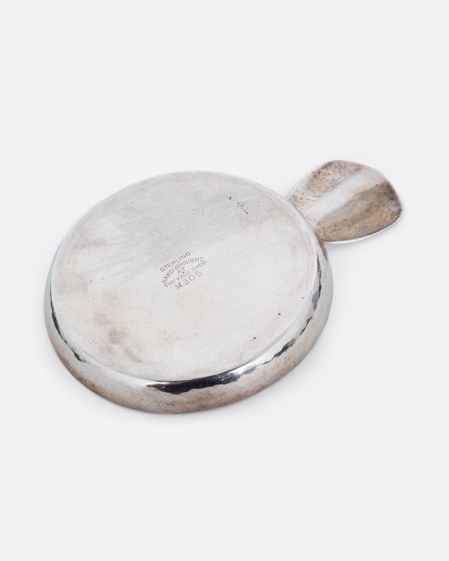 A round silver ashtray with a ledge for placing a cigarette. Shown from the back at an angle.