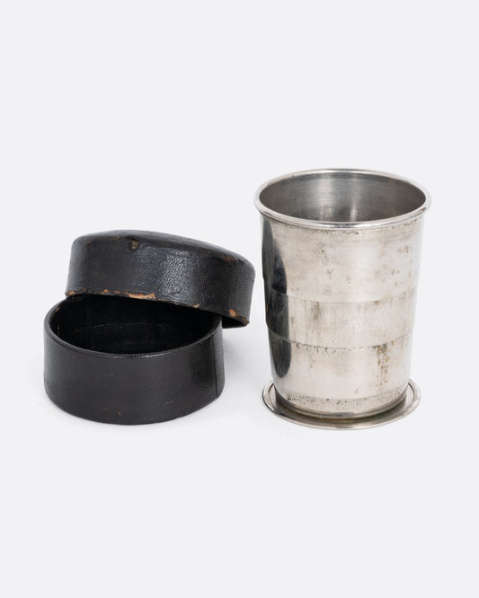A silver  collapsible cup with a round leather case open to its side. Shown from the front.