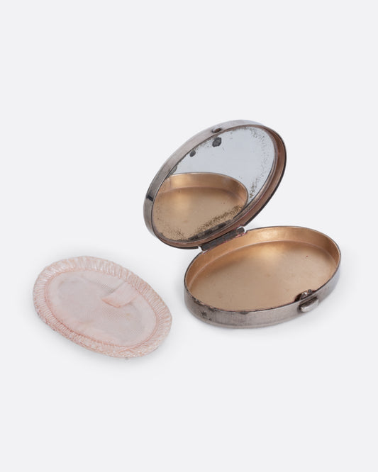A silver mirror compact shown open at an angle from the front with a powder puff sitting at its side.