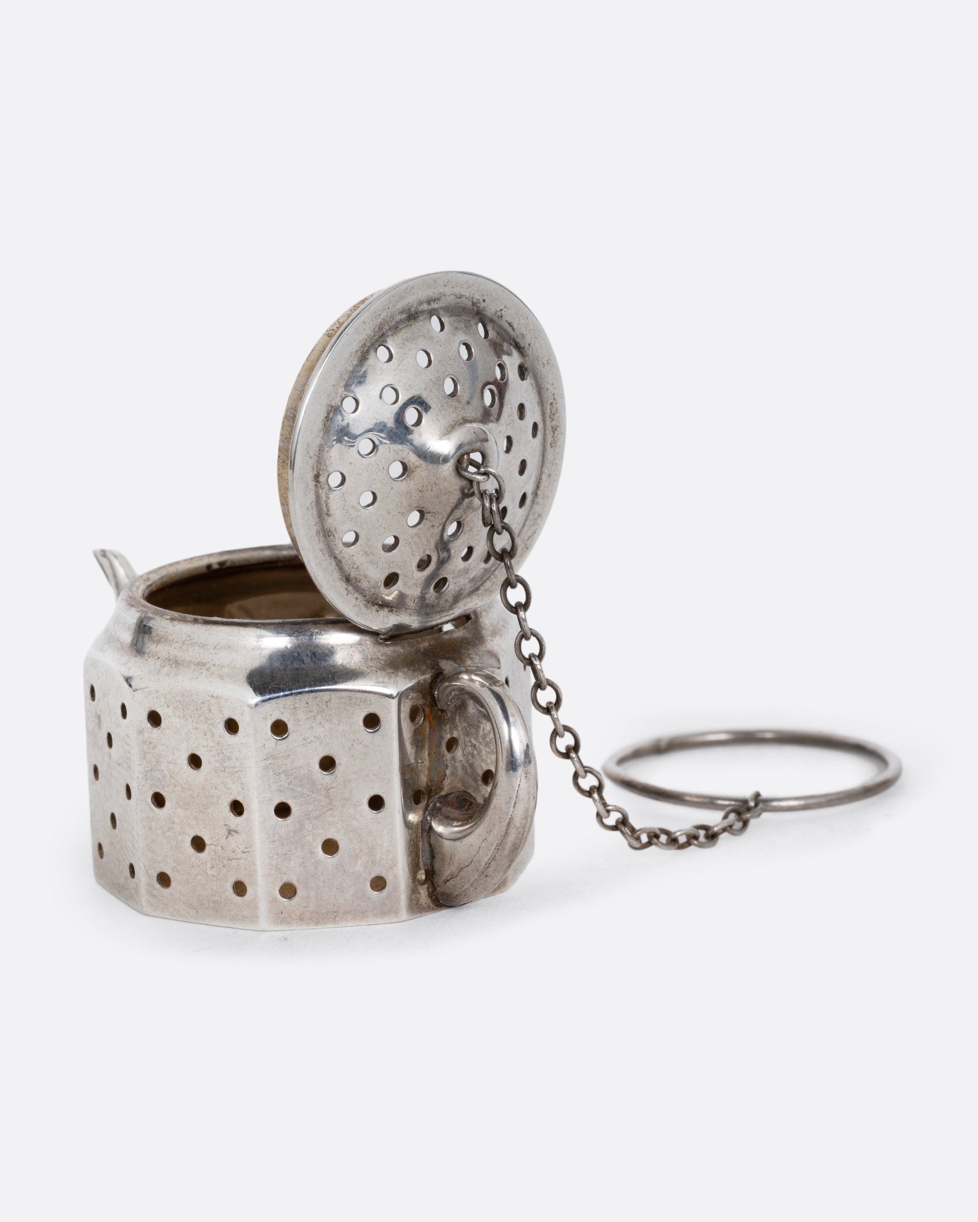 A silver teapot shaped tea infuser with a chain and hoop attached to the lid. Its hinged lid is shown open. Shown at an angle from the back.
