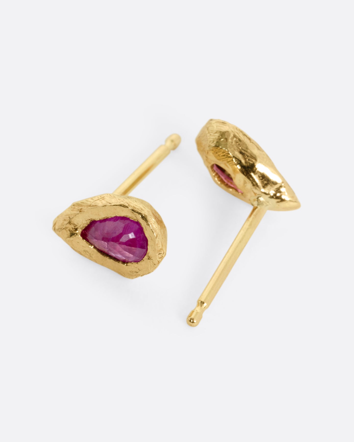 A pair of yellow gold stud earrings with teardrop shaped rubies, shown lying on their sides; one earring from the front, the other from the back.
