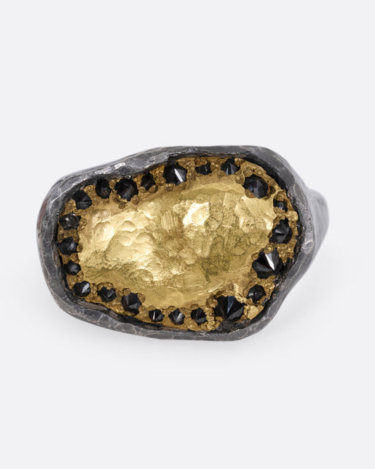 A white gold signet ring with a yellow gold face and a halo of reverse set black diamonds. Shown from the front.