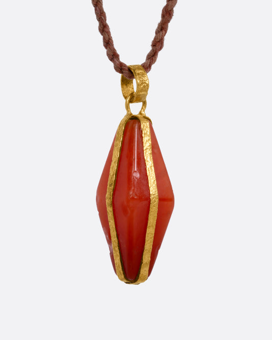 A carnelian pendant wrapped in yellow gold and suspended from a cord, shown from the side. 