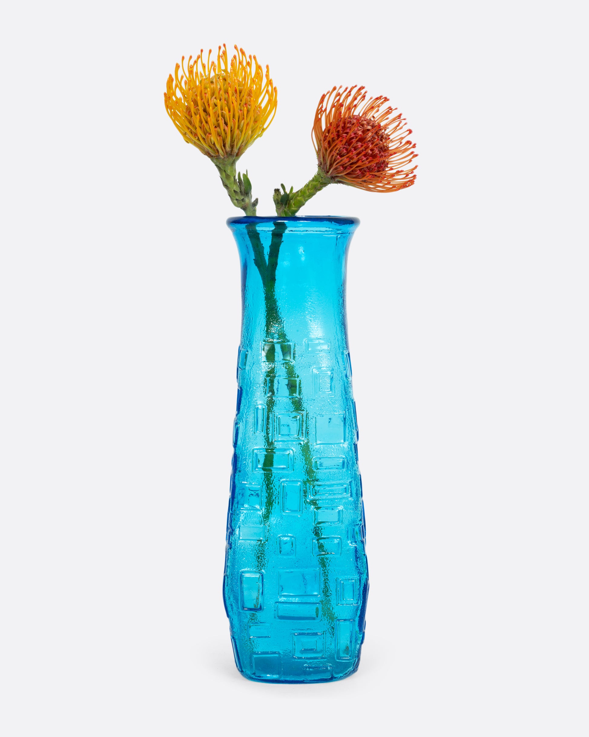 A tall cerulean glass vase with an all-over geometric pattern. Shown with orange and yellow flowers.