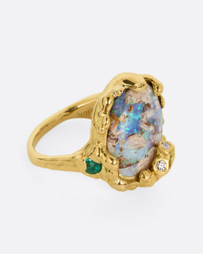 A yellow gold ring with a fossilized wood opal and diamond and emerald accents, shown from the side.
