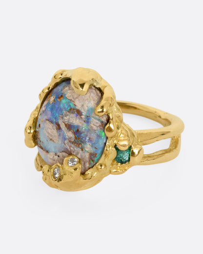 A yellow gold ring with a fossilized wood opal and diamond and emerald accents, shown from the side.