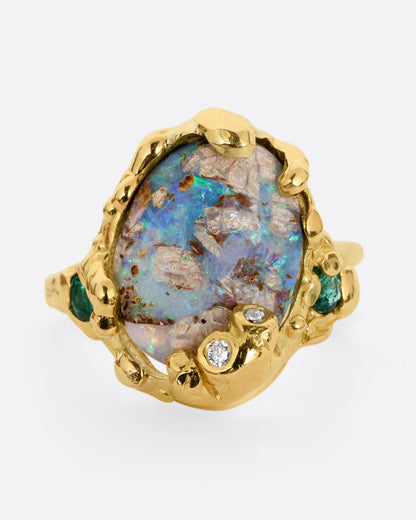 A yellow gold ring with a fossilized wood opal and diamond and emerald accents, shown from the front.