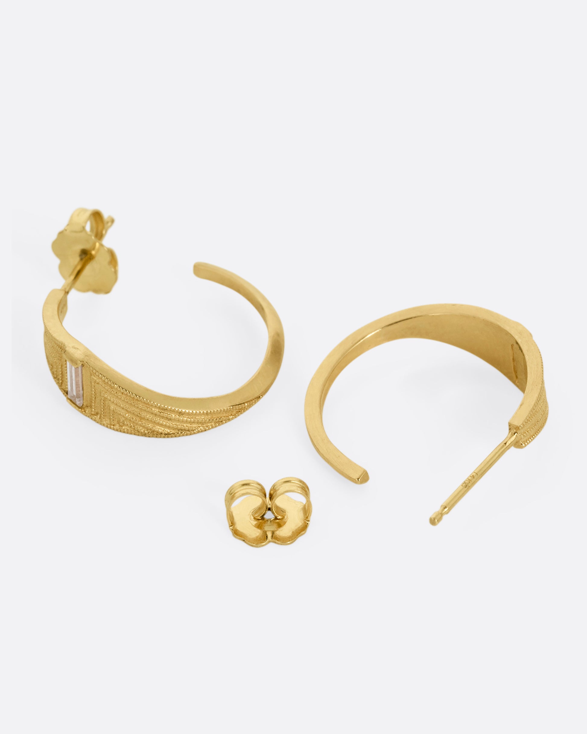 A pair of yellow gold deco inspired hoops with a baguette diamond and a geometric pattern. Shown lying flat; one earring from the front, the other open from the back.