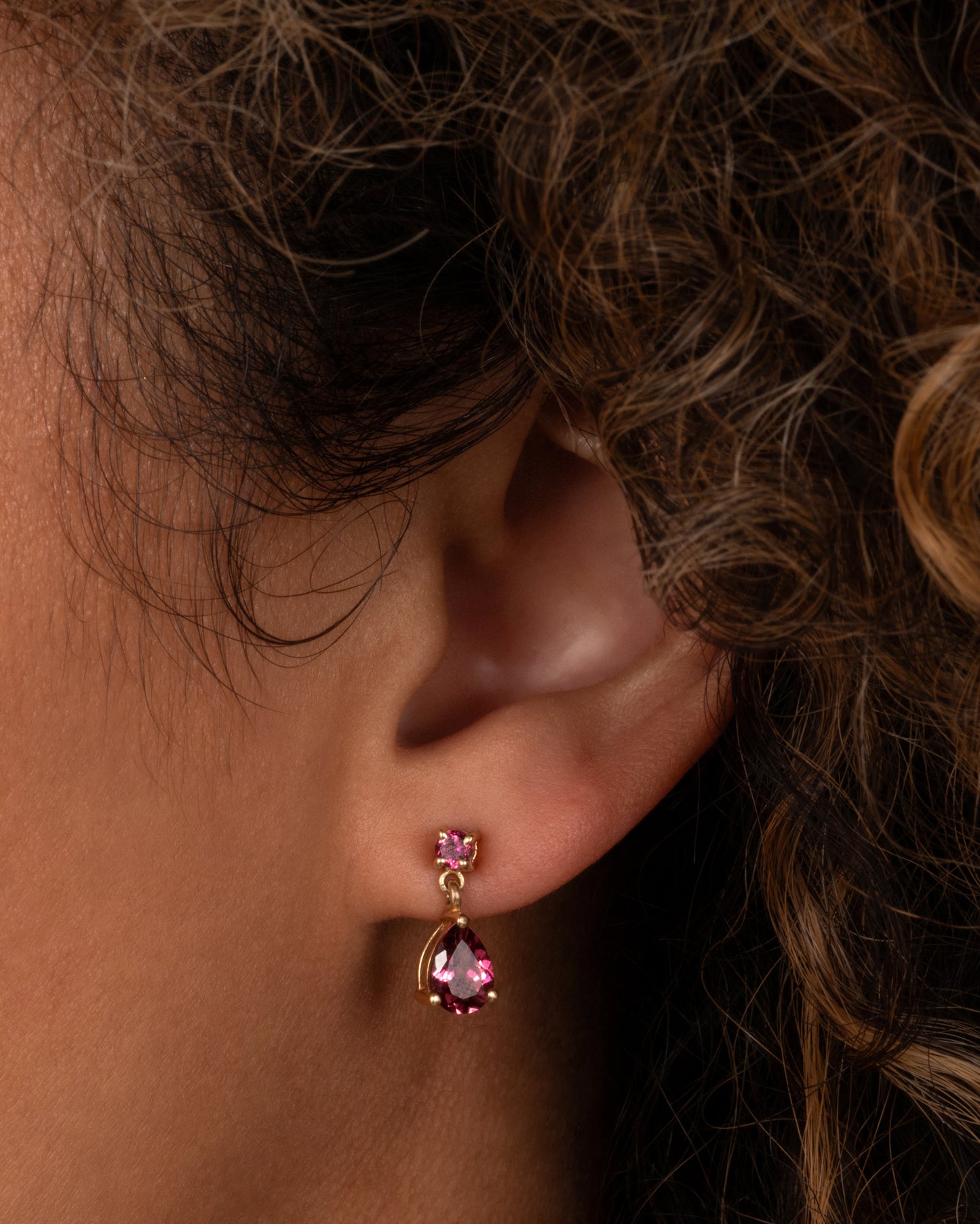 Yellow gold earrings with round prong set pink tourmalines at the top and teardrop shaped pink tourmalines at the bottom. Shown worn in an ear lobe.