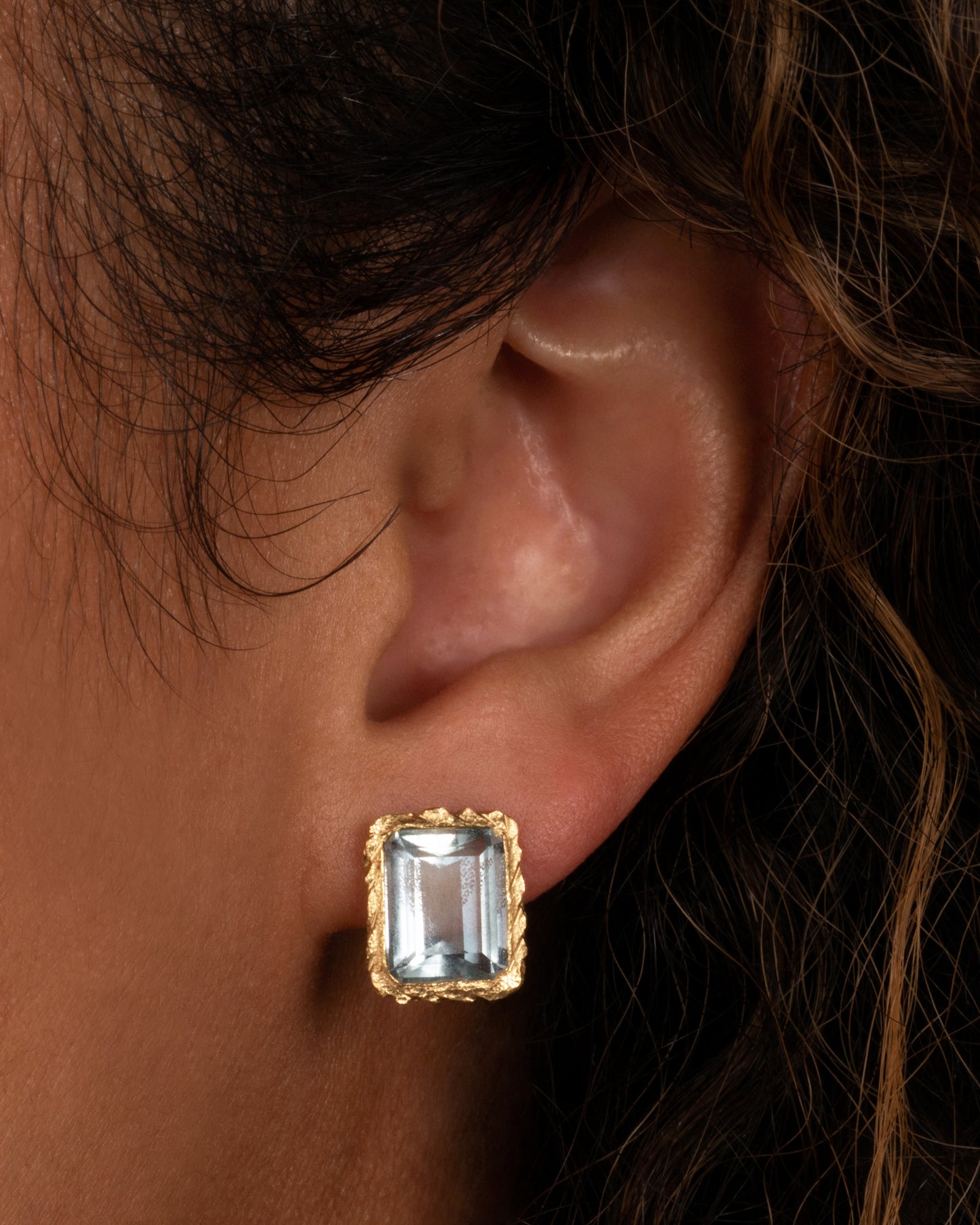 A yellow gold stud earrings with rectangular aquamarines. Shown on an ear. 