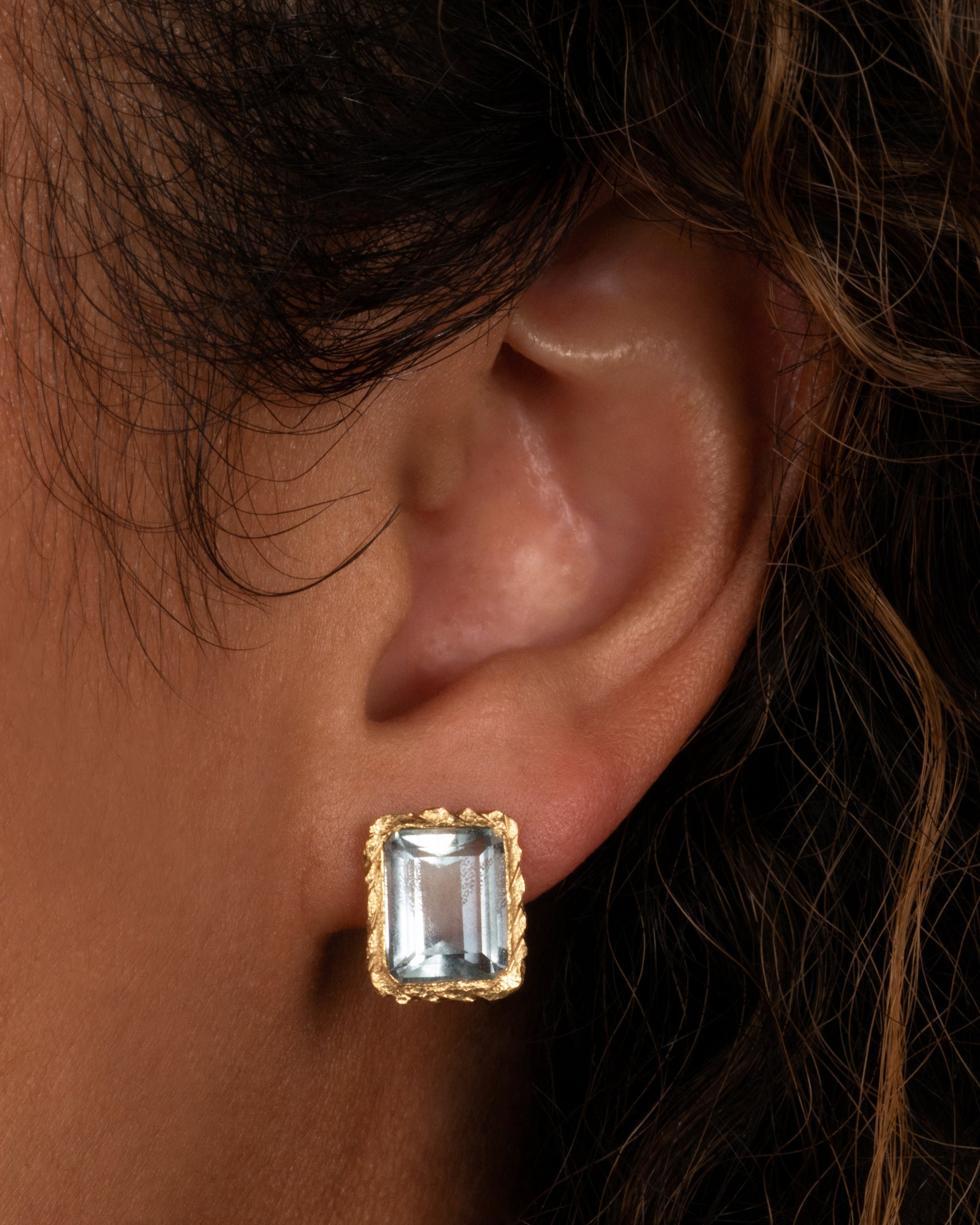A yellow gold stud earrings with rectangular aquamarines. Shown on an ear. 