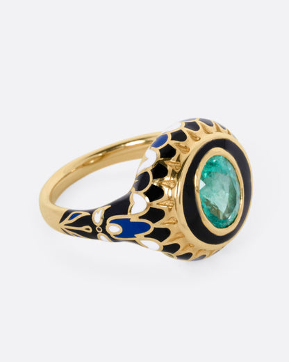 A yellow gold ring with an intricate enamel pattern around a bezel set oval emerald, shown from the side.