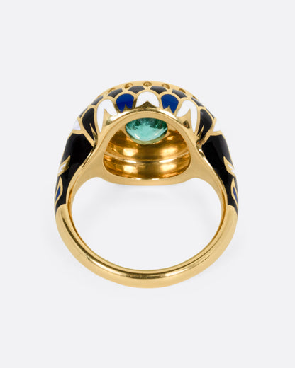 A yellow gold ring with an intricate enamel pattern around a bezel set oval emerald, shown from the back.
