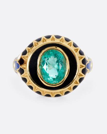 A yellow gold ring with an intricate enamel pattern around a bezel set oval emerald, shown from the front.