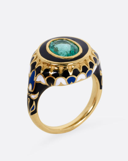 A yellow gold ring with an intricate enamel pattern around a bezel set oval emerald, shown from the side standing up.