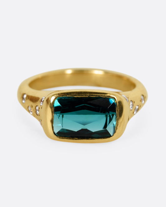 A yellow gold ring with a rectangular green tourmaline at the center and diamonds on either shoulder. Shown from the front. 