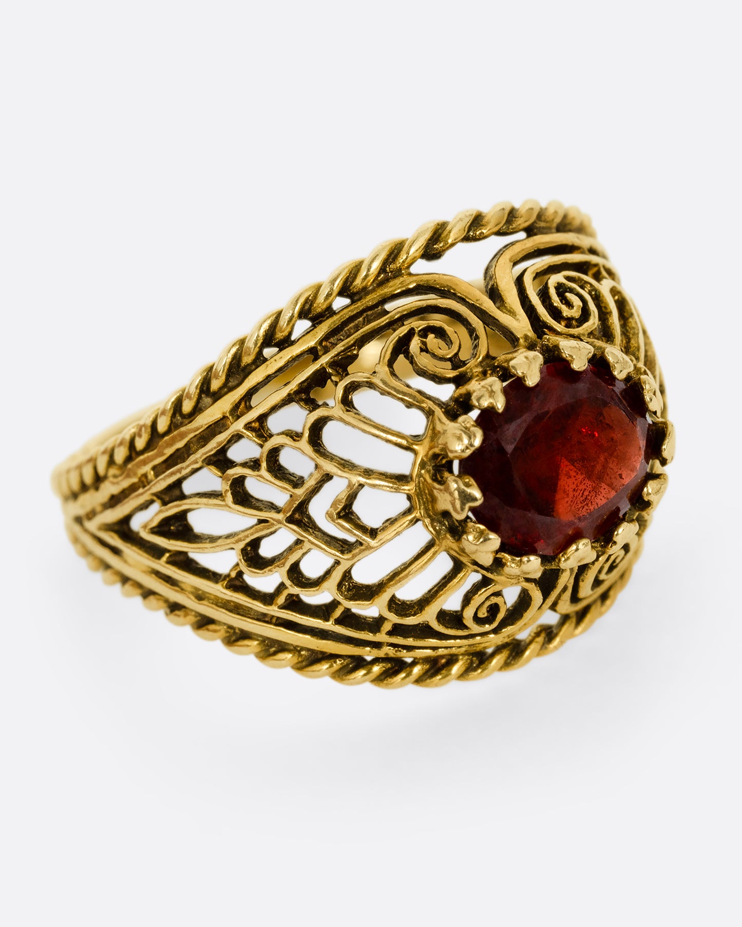 A yellow gold dome ring with an intricate web of filigree and delicate cutouts and a garnet at its center. Shown from the side.