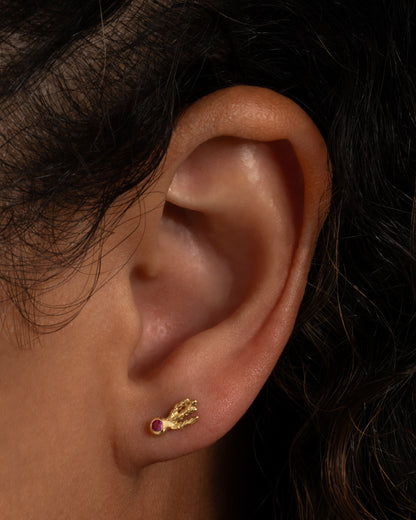 A yellow gold stud earring with a pink sapphire and comet-like tails. Shown worn on an ear.