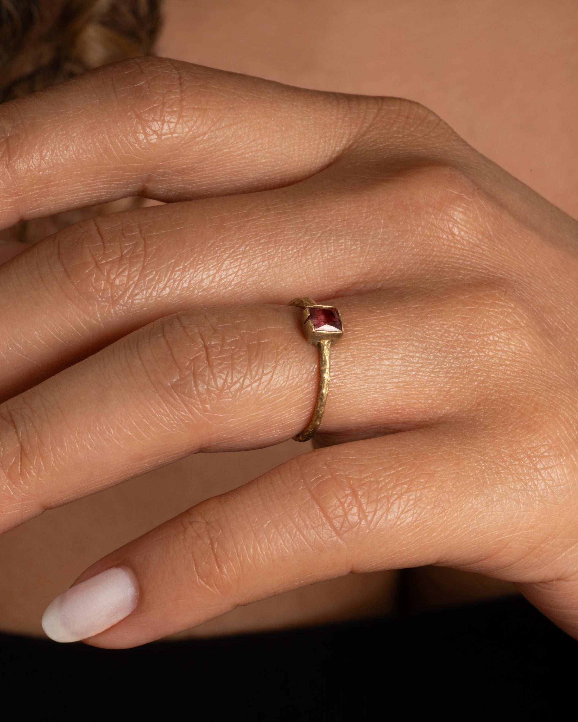 A textured yellow gold ring with a square pink tourmaline. Shown on a finger.