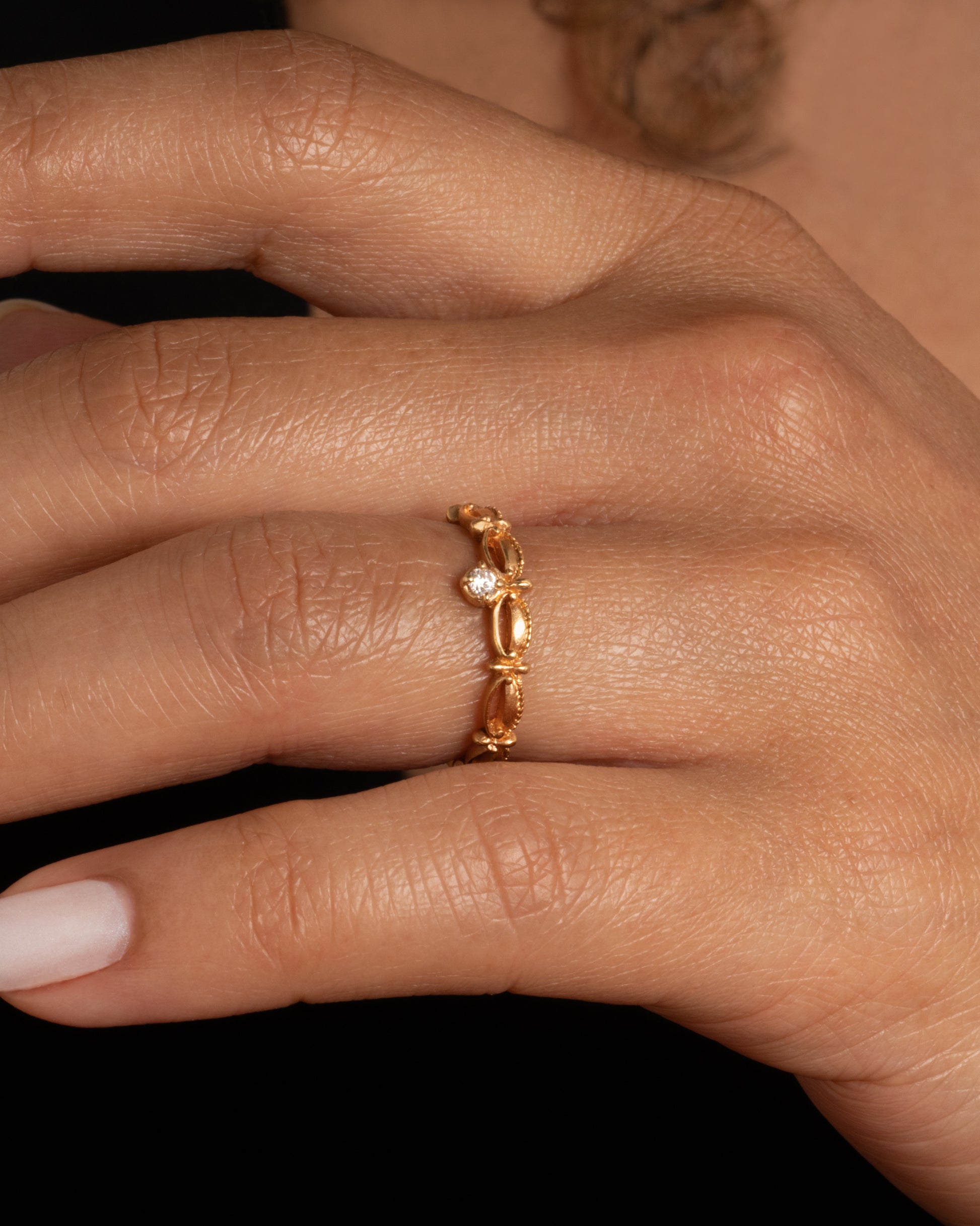 A yellow gold tiara shaped band with curved and textured details and a round white diamond at the center. Shown worn on a finger.