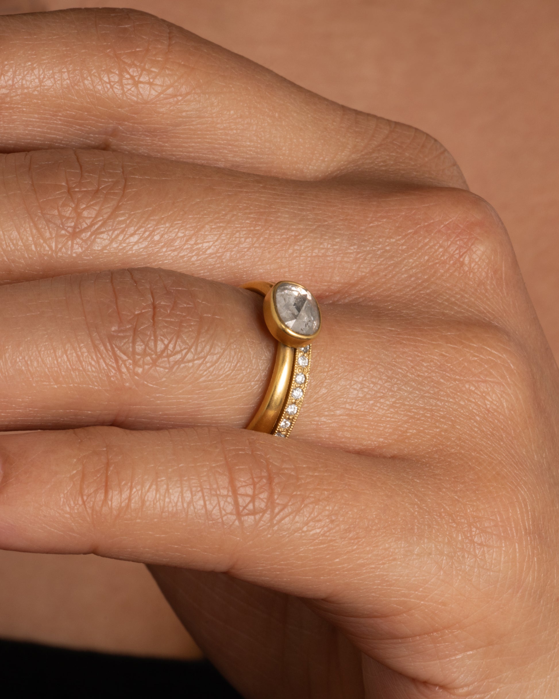 A pale grey, rose cut, oval diamond. Set horizontally on a simple, yellow gold band. Shown worn on a finger paired with a Todd Pownell bead set diamond eternity band.