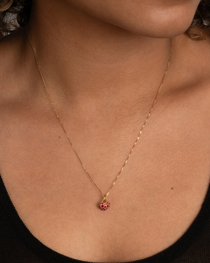 A yellow gold heart shaped charm with inverted rubies throughout. Shown worn on a chain around the neck.