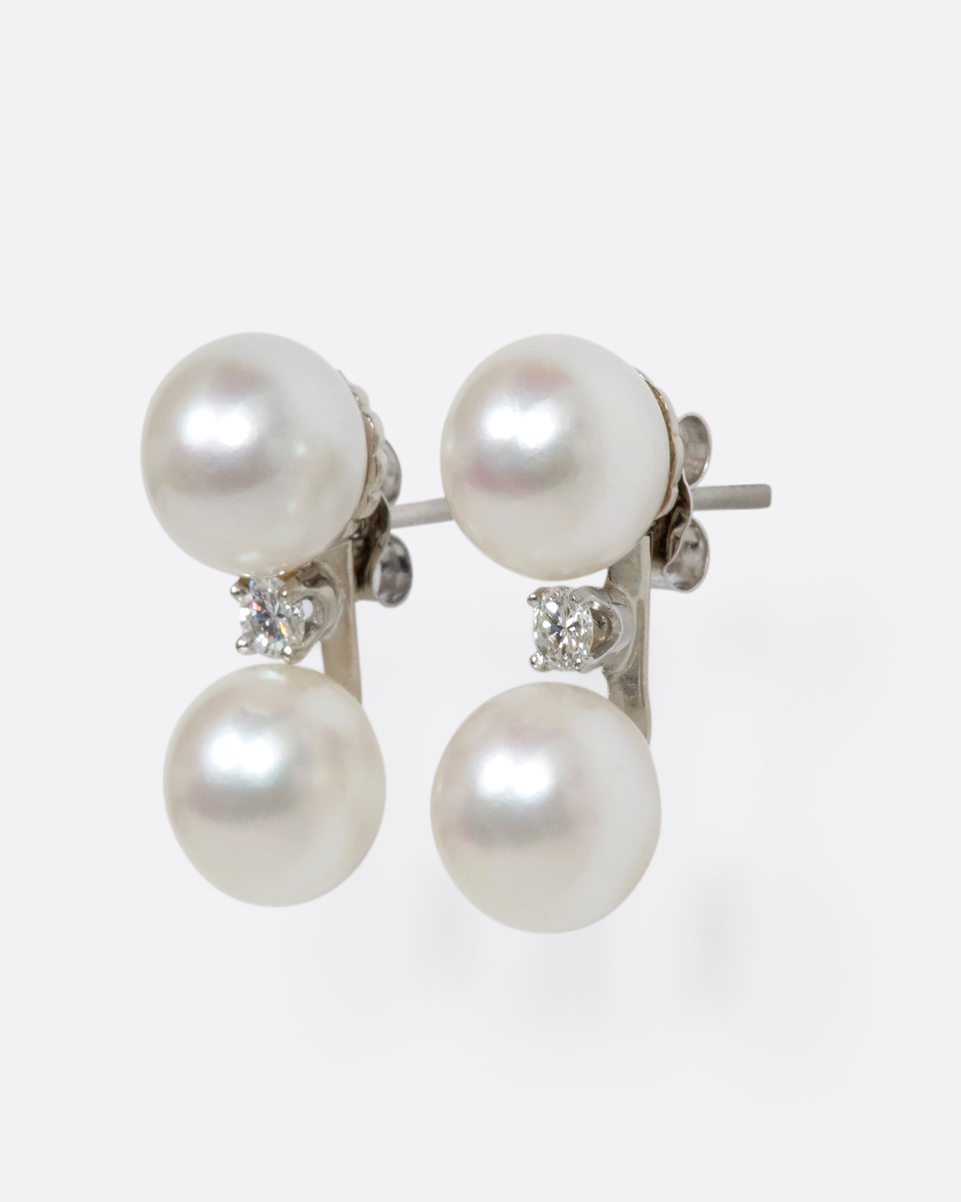 A pair of white gold studs featuring two pearls with a diamond situated in between. View from the side.