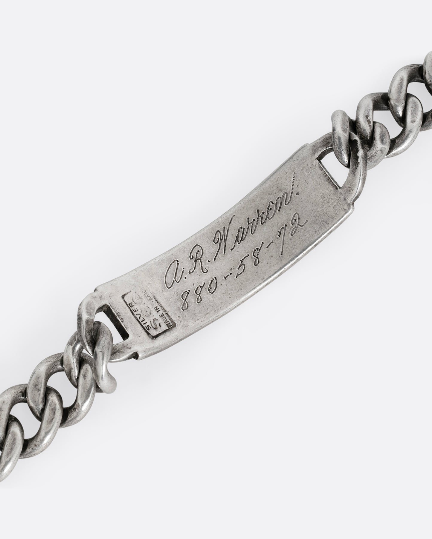 A close up of a silver cable chain ID bracelet with "A.R. Warren" in curling script  engraved on the inside of its name tag. 