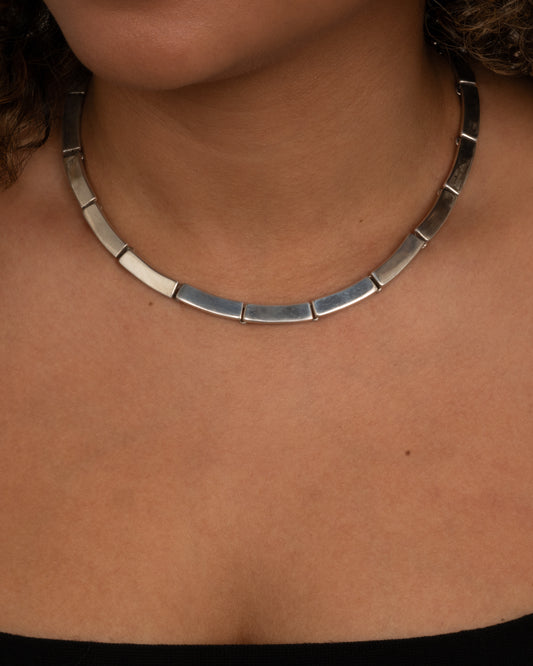 A silver necklace with hinged rectangular links shown from the front on a person.