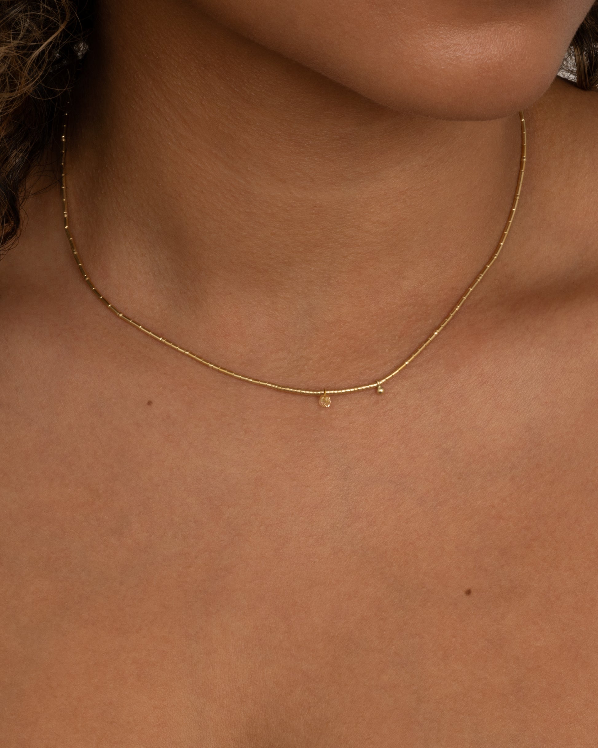 A yellow gold beaded necklace with a diamond briolette and pops of red cording beside the clasp. Shown from the front on a person.