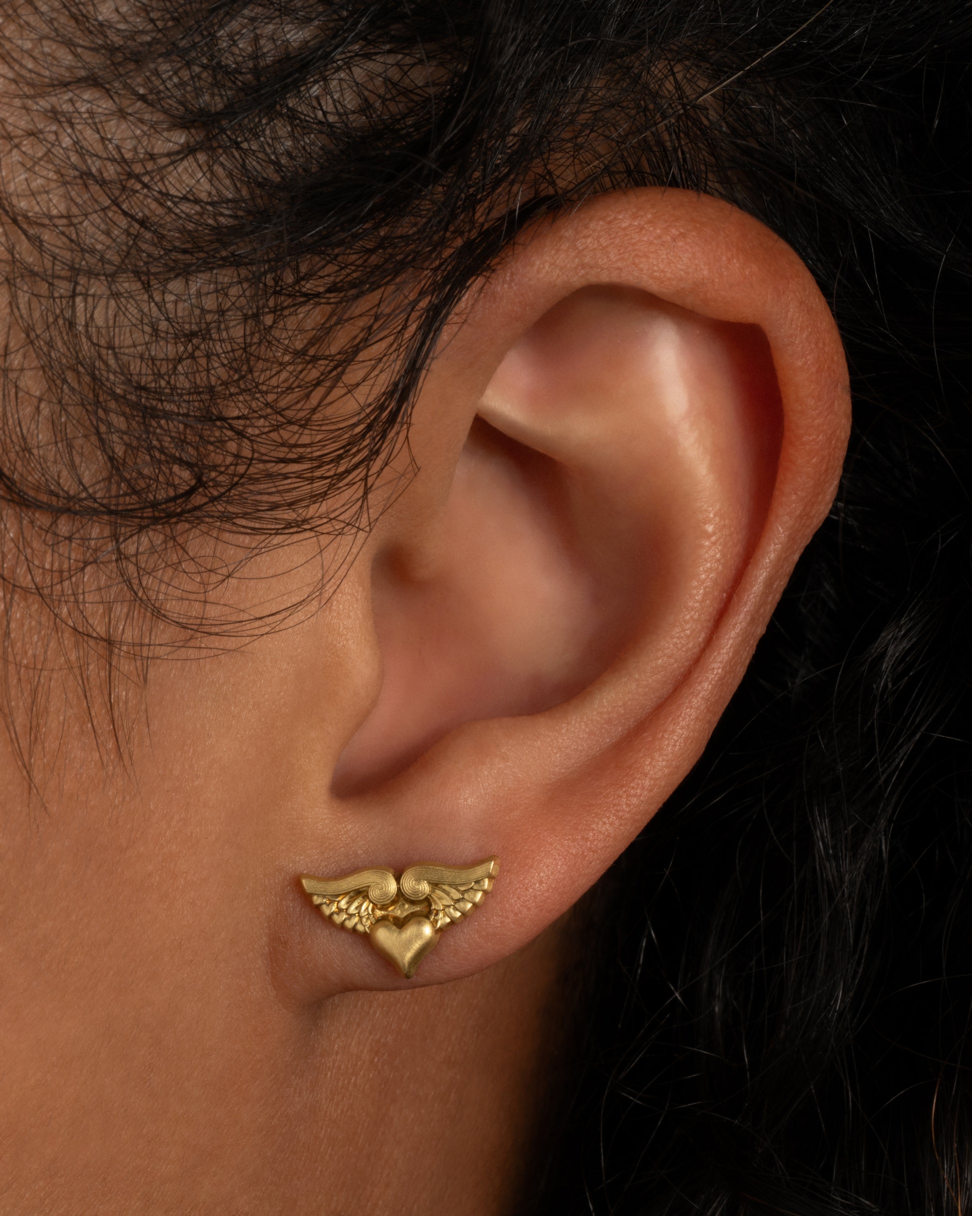 A yellow gold stud earring with a heart with wings. Shown from the front on a person.