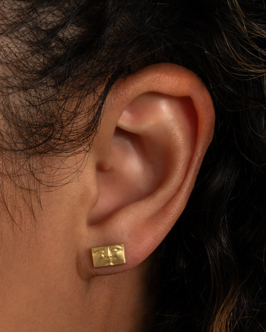 A yellow gold rectangular brick stud earring with a face. Shown worn in an ear.