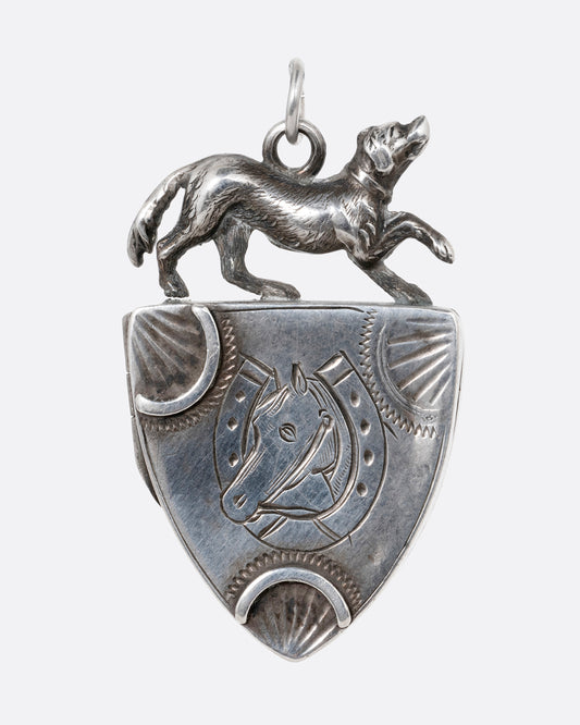 A silver shield- shaped locket with a dog perching on top and an engraved horseshoe framed mare. Shown from the front.