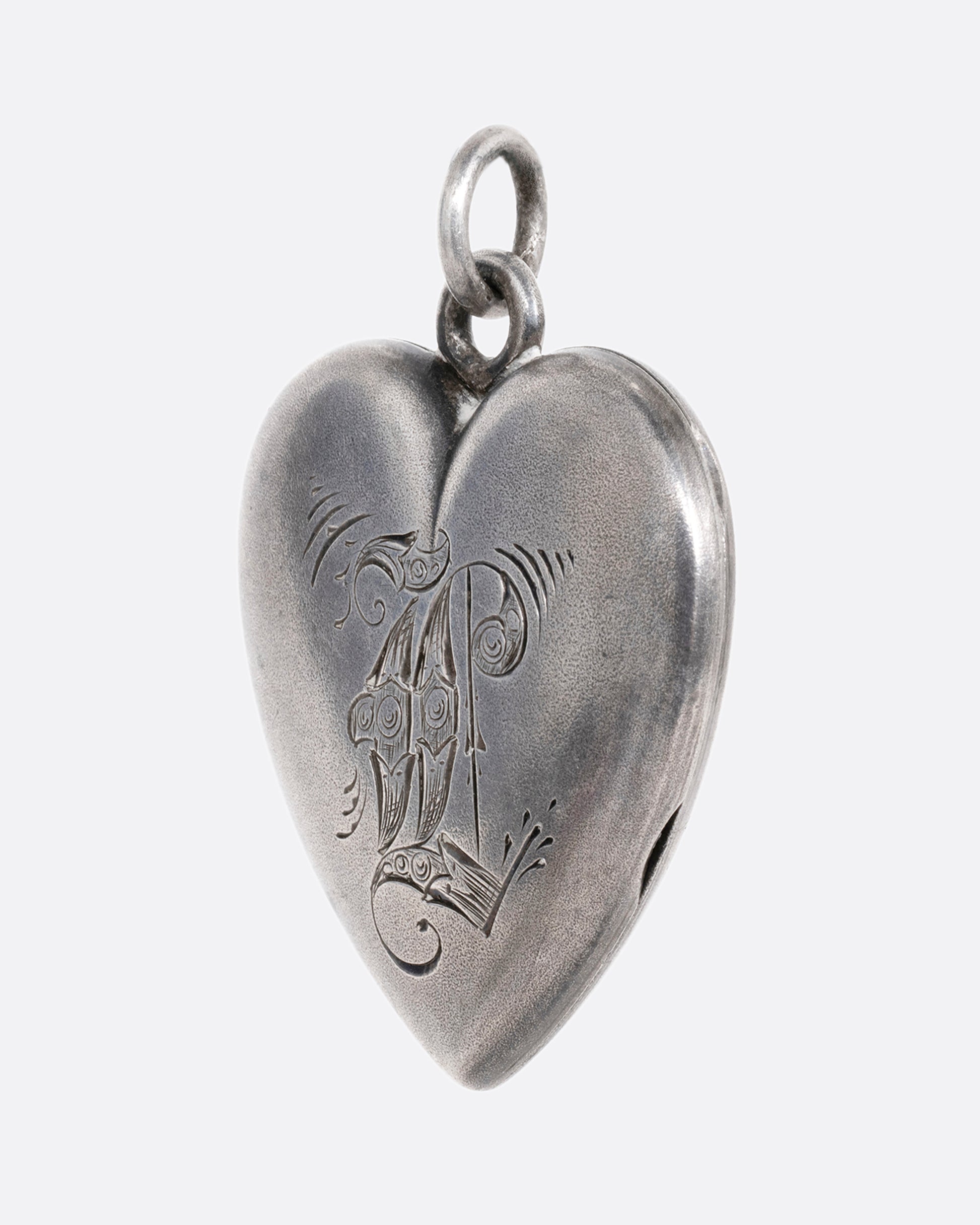 A silver heart shaped locket with an engraved 'L'. Shown from the side. 