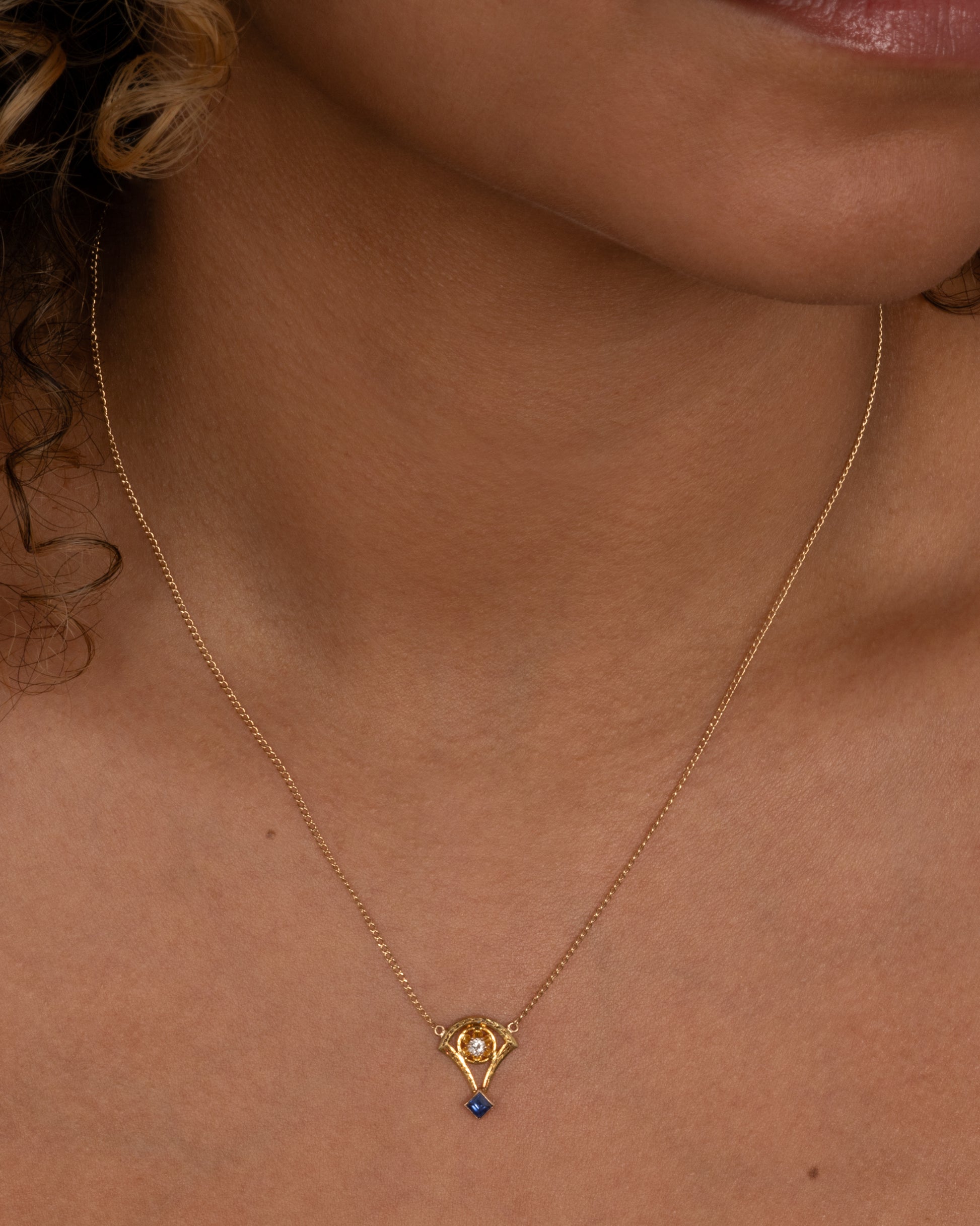 A yellow gold cable chain necklace with a pendant shaped like the basket of a mini hot air balloon with a prong set diamond. and princess cut sapphire. Shown from the front on a person.