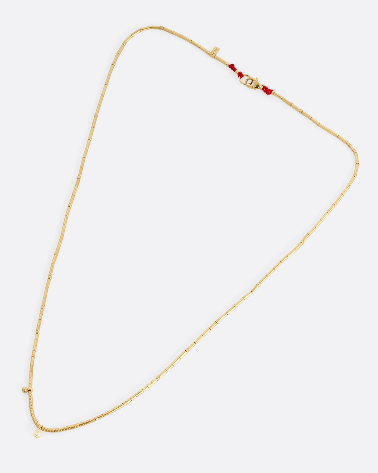 A yellow gold beaded necklace with a diamond briolette and pops of red cording beside the clasp. Shown lying flat.