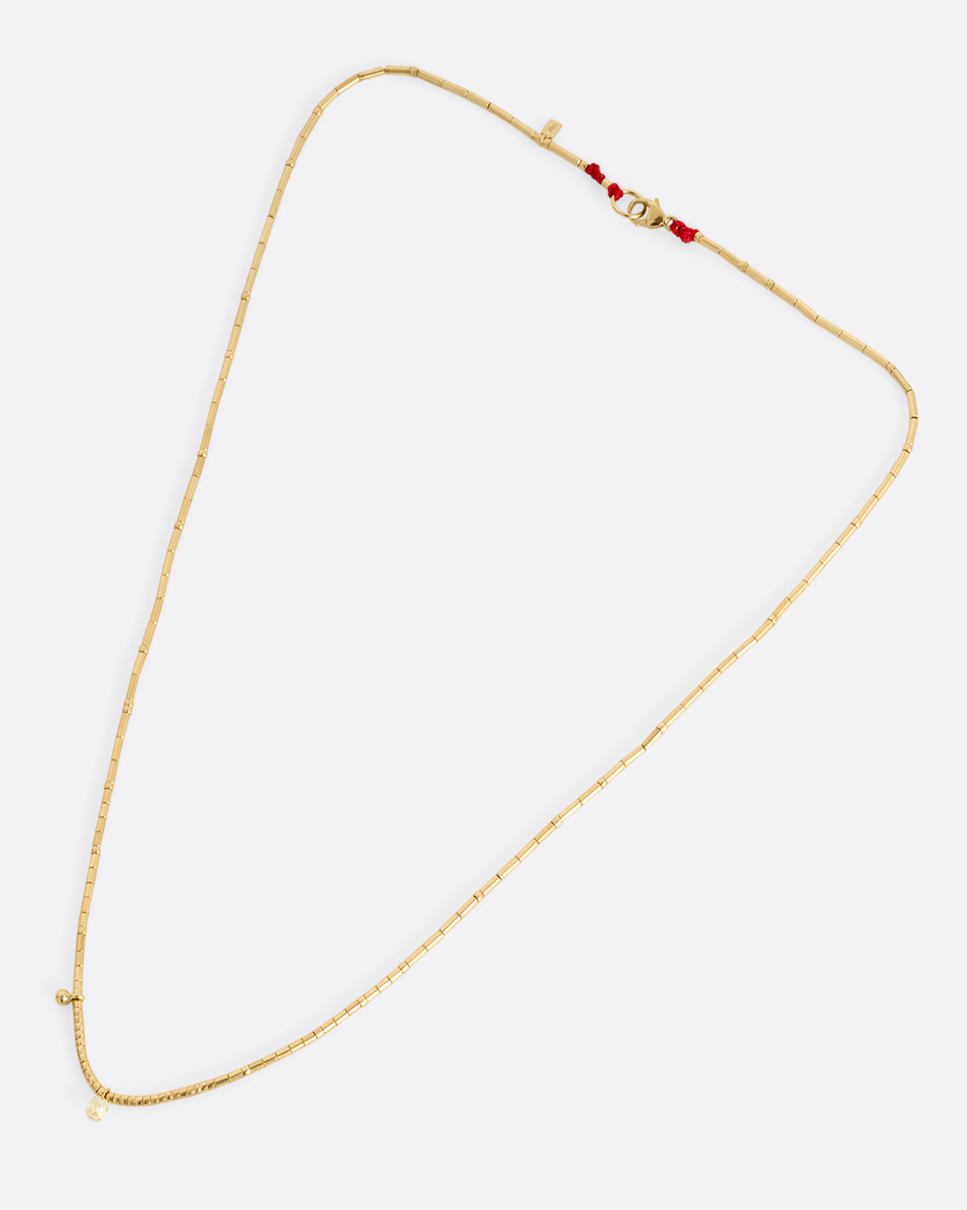 A yellow gold beaded necklace with a diamond briolette and pops of red cording beside the clasp. Shown lying flat.