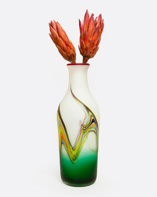 An opaque handblown vase with a colorful swirled motif and red rim, shown from the front with two red protea flowers in it.