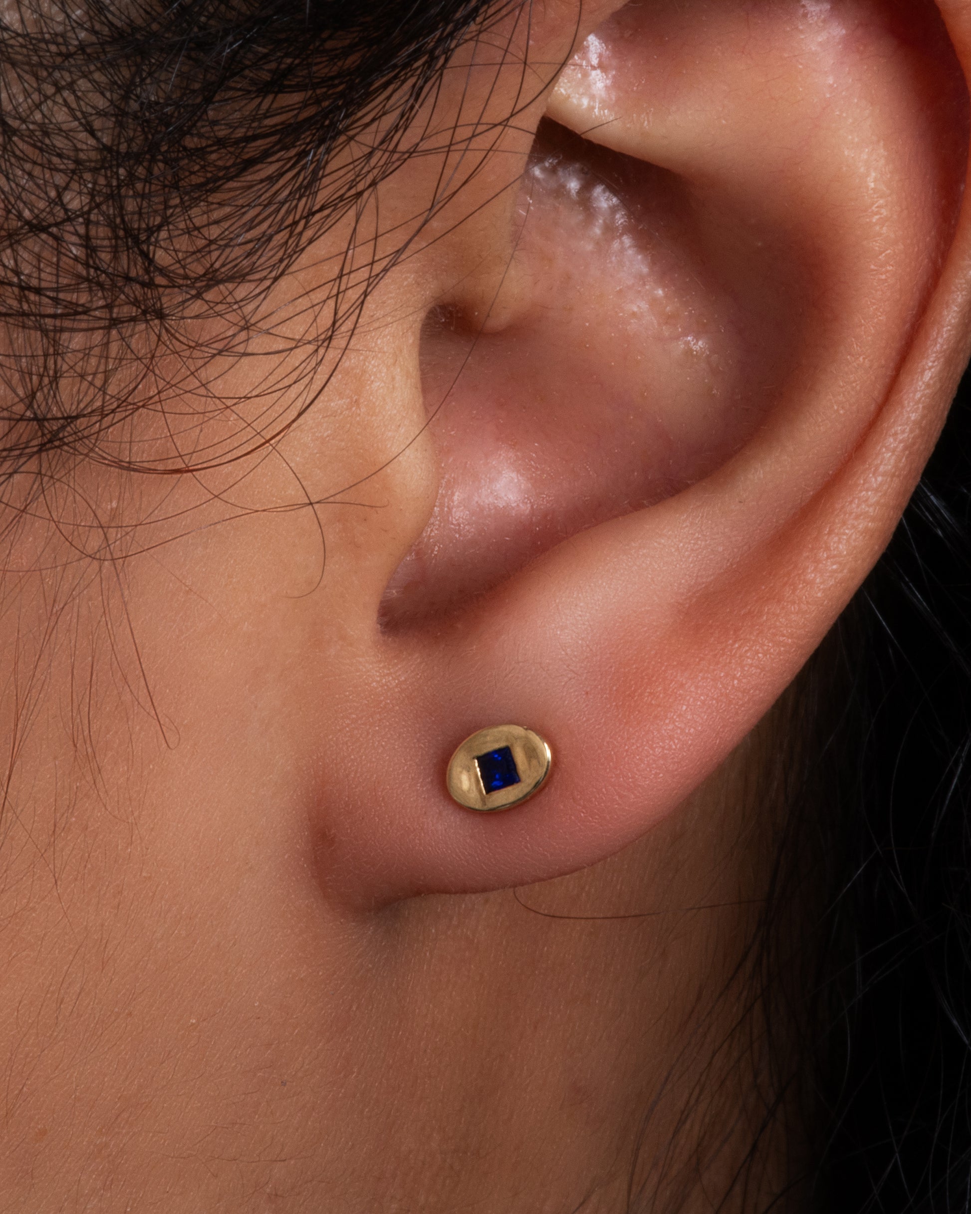 A pair of oval yellow gold stud earrings, each with a princess cut blue sapphire at its center. Shown on an ear.