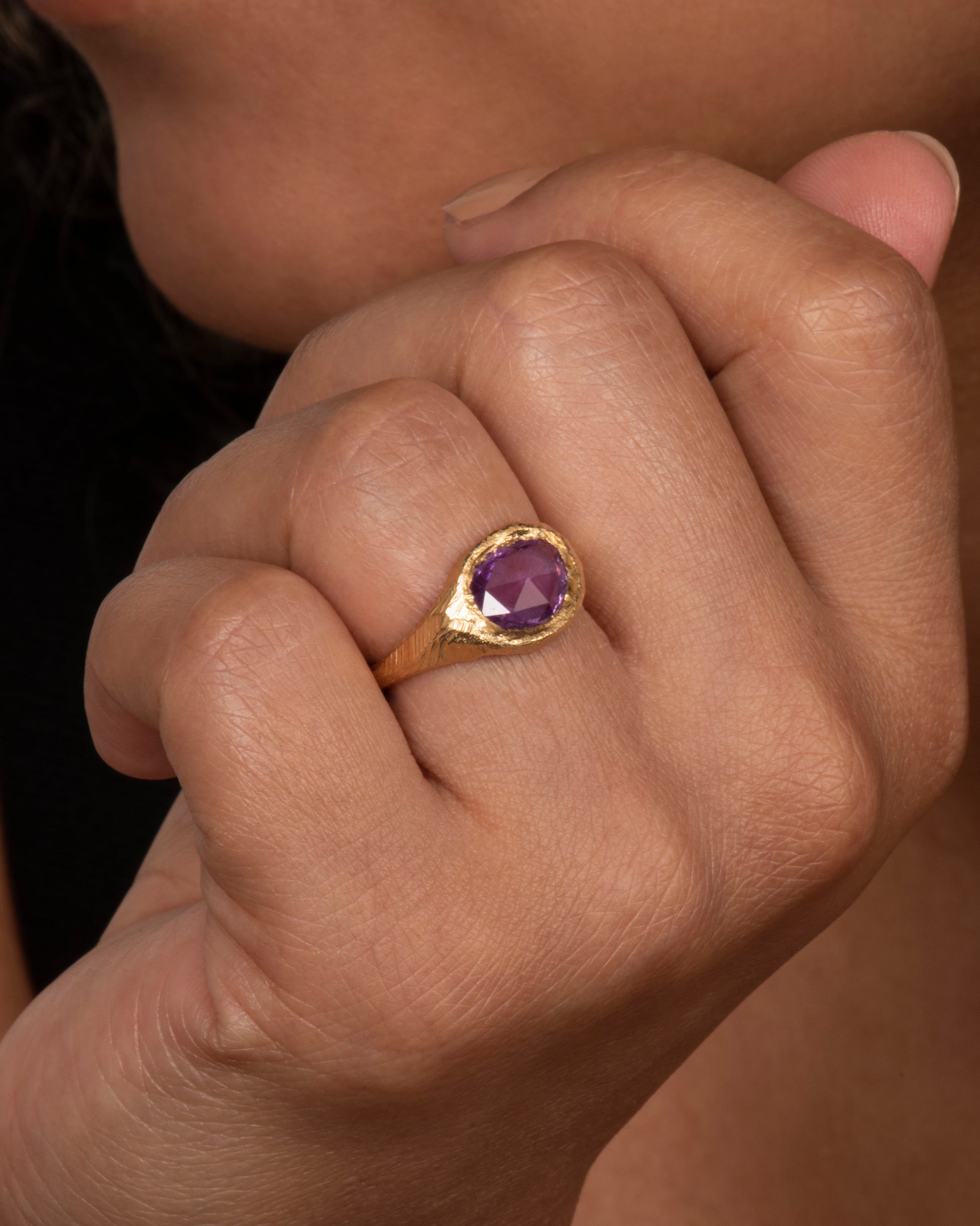 A textured yellow gold signet style ring with a rose cut purple sapphire. View on a hand.