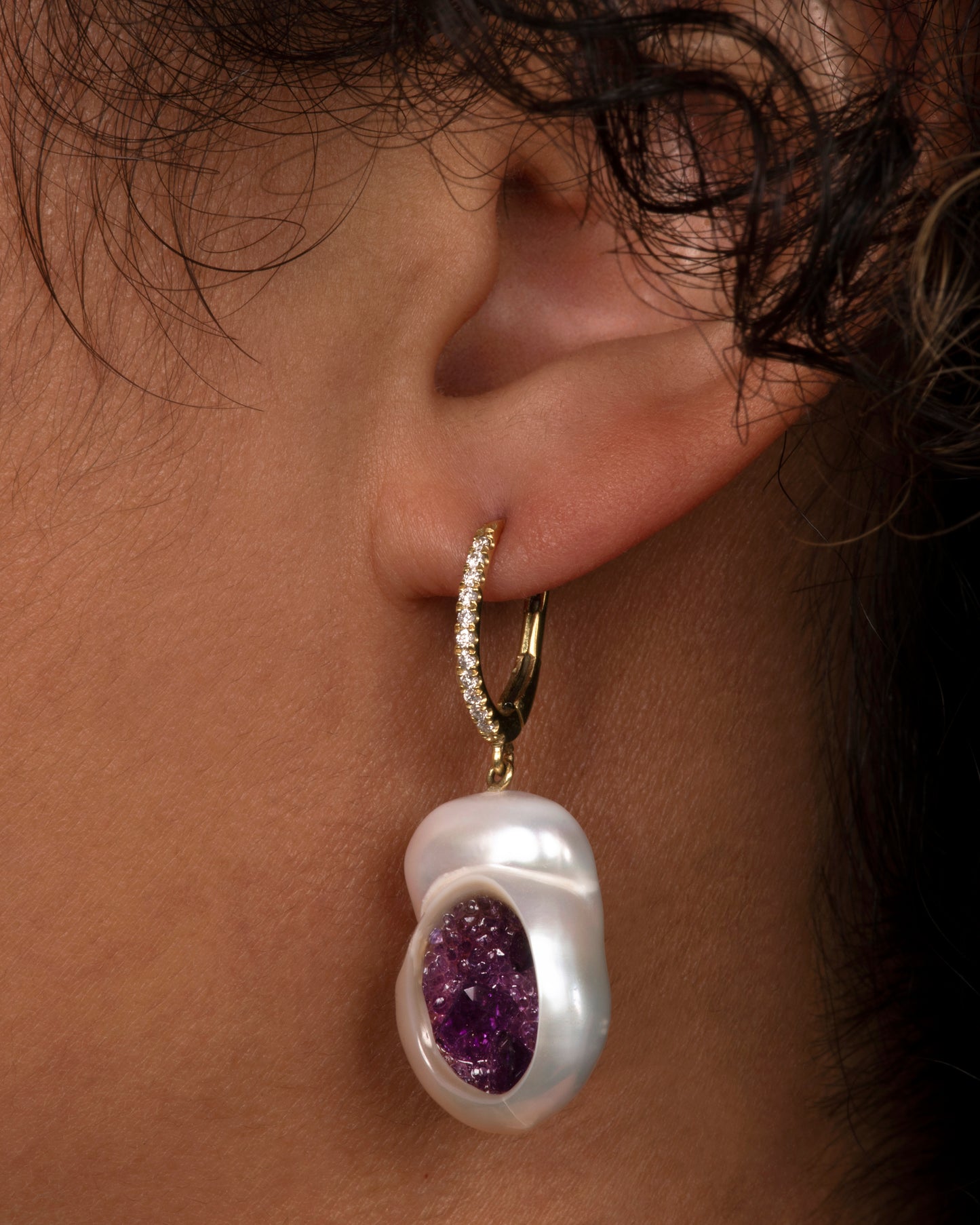 two dangle drop earrings hanging and viewed from the side. the drop is a pearl that has been partially carved out and lined with amethysts. the yellow gold hooks have pave diamonds on the bail. Shown on an ear.