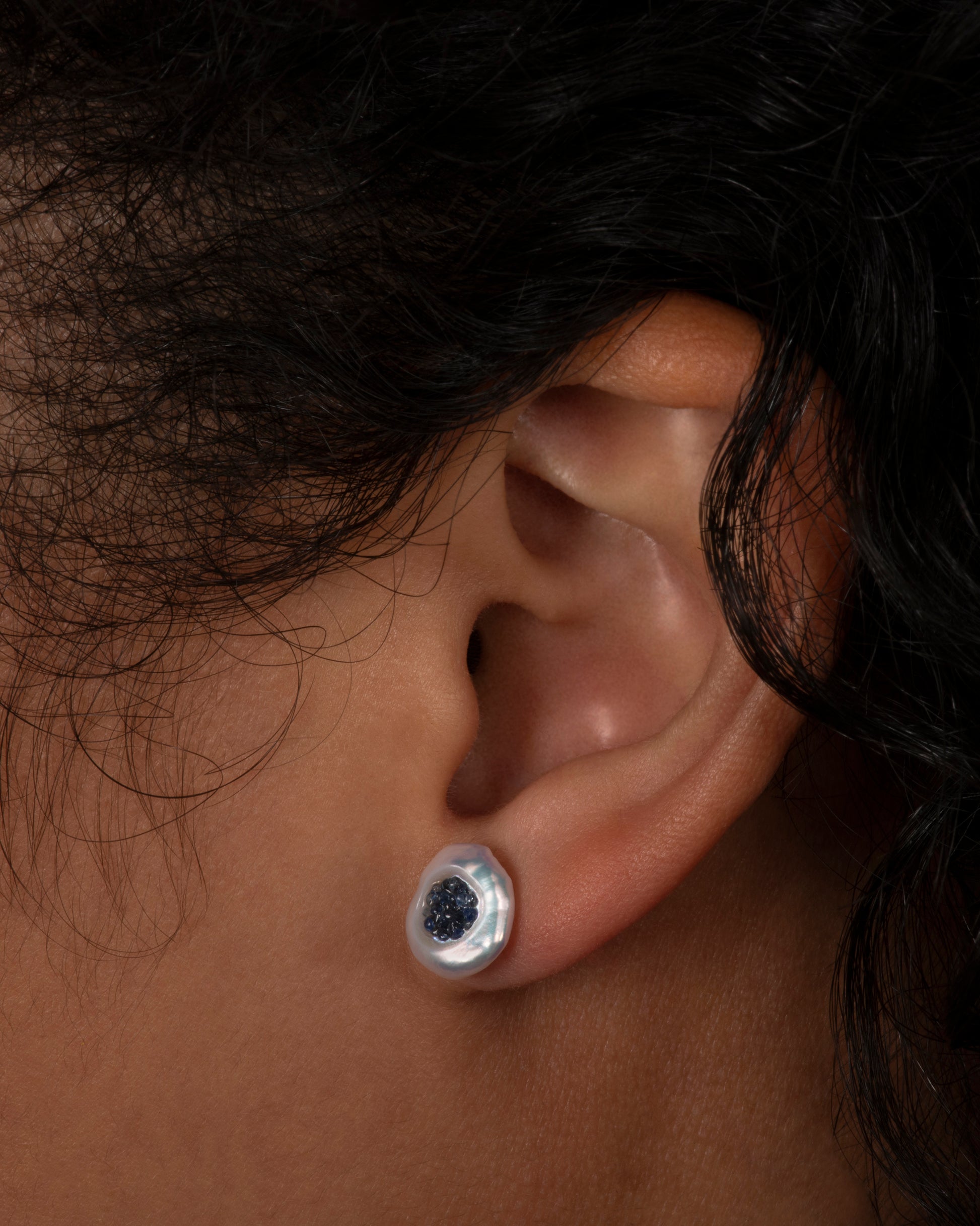 A pair of yellow gold earrings with hollowed out pearls filled with sapphires. Shown on an ear.