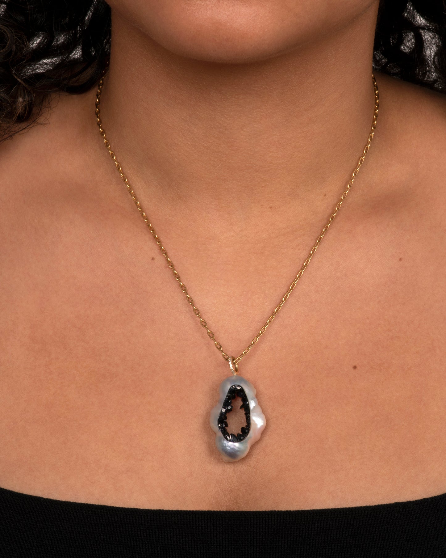 A yellow gold necklace with a hollowed souffle pearl pendant filled with a blue sapphire geode hanging from a pave diamond bale. Shown worn around the neck.