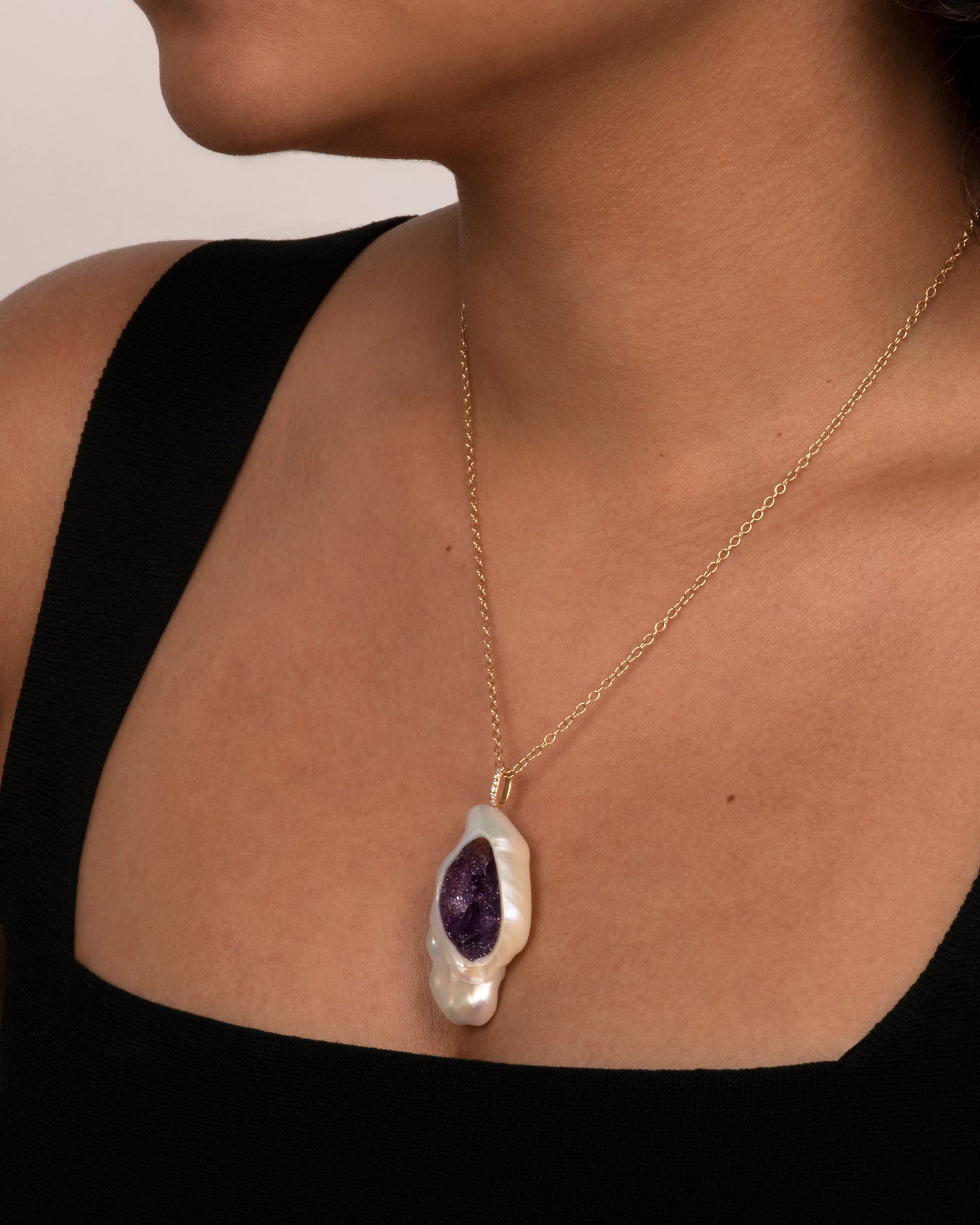 A yellow gold necklace with a hollowed out pearl pendant filled with amethysts and a pave diamond bale. Shown on a neck.