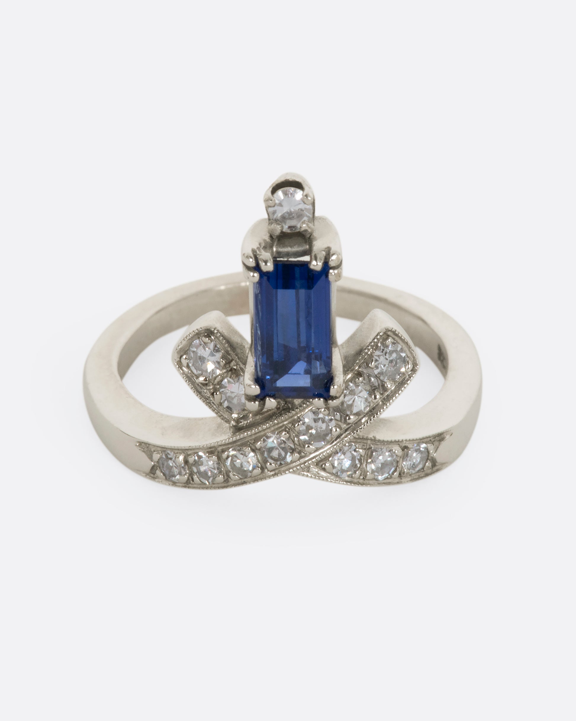 A white gold crossed band ring with a baguette blue sapphire topped with a diamond. View from the front slightly above.