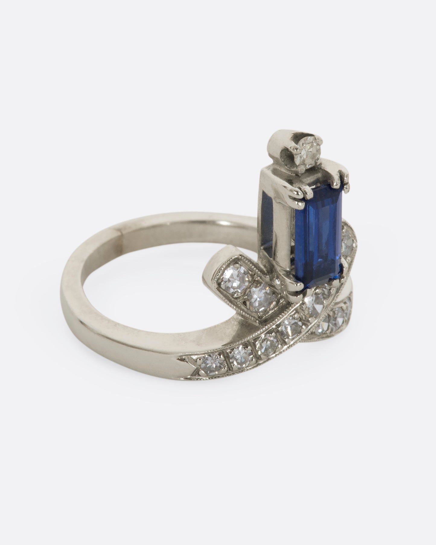 A white gold crossed band ring with a baguette blue sapphire topped with a diamond. View from the alternate side.