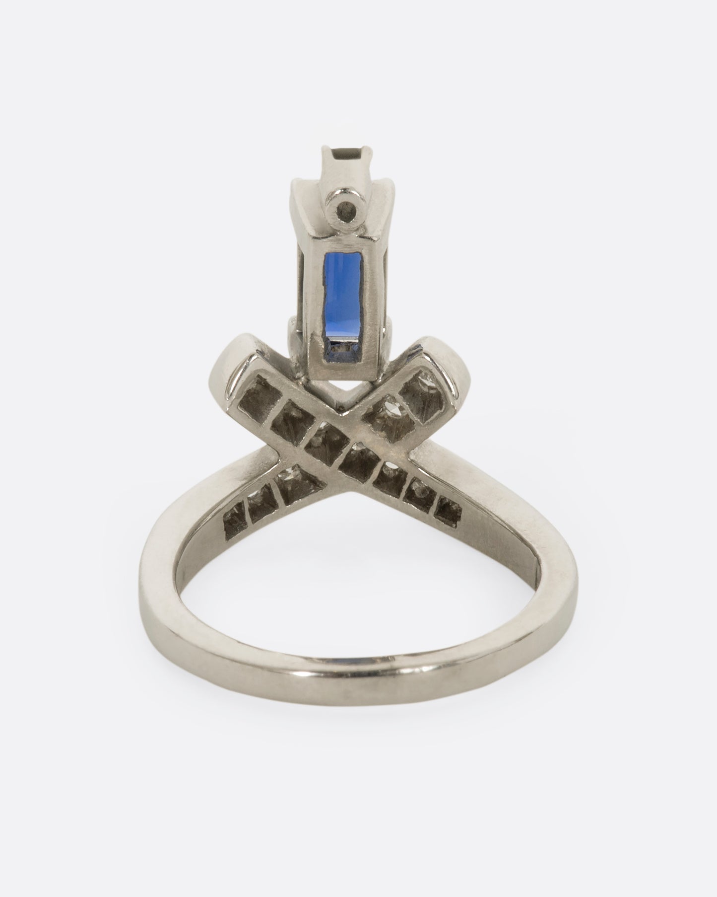 A white gold crossed band ring with a baguette blue sapphire topped with a diamond. View from the back.