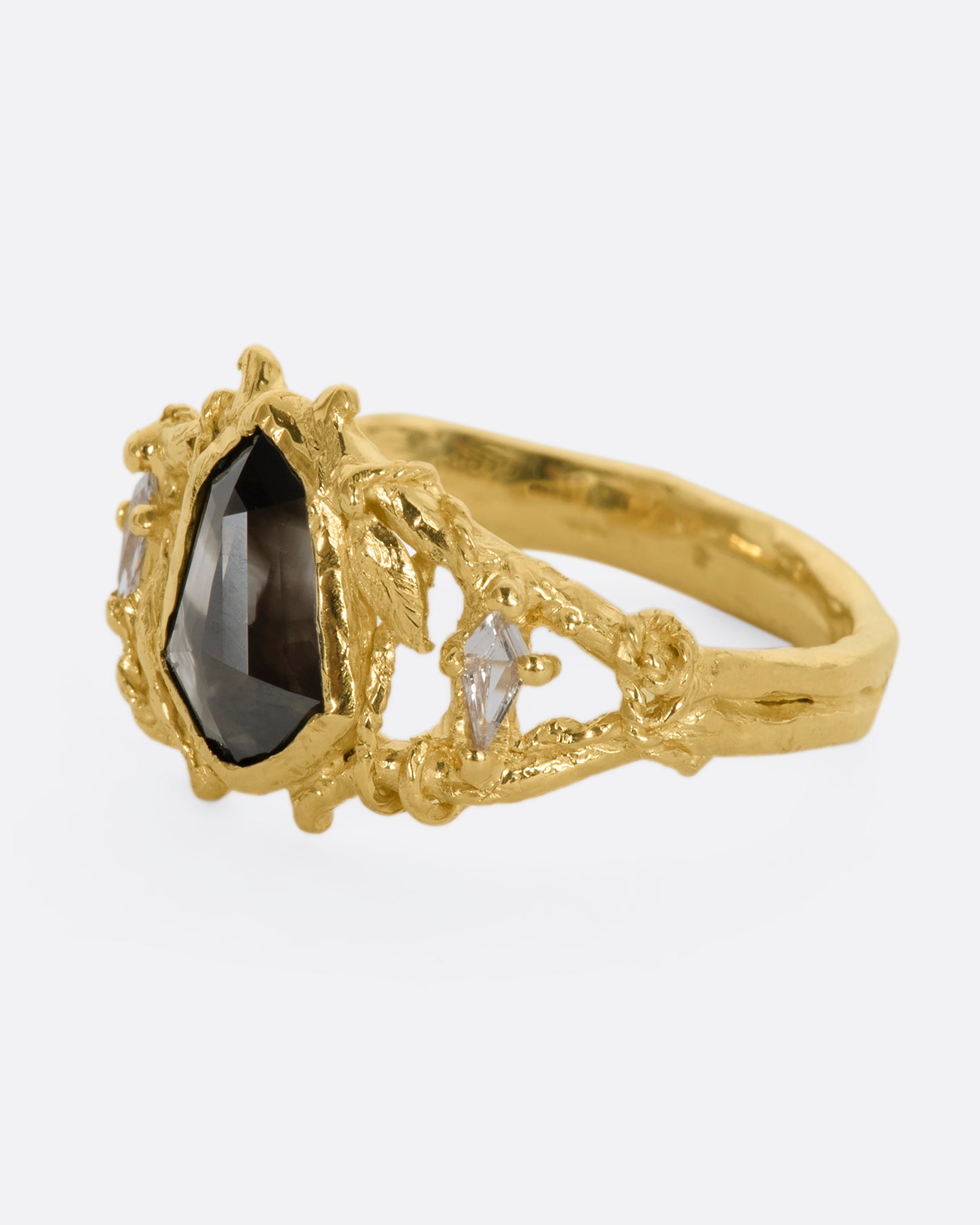 A yellow gold ring with an organic vine motif and a shield double cut black diamond at the center with white kite sapphires on either side. View from the side.