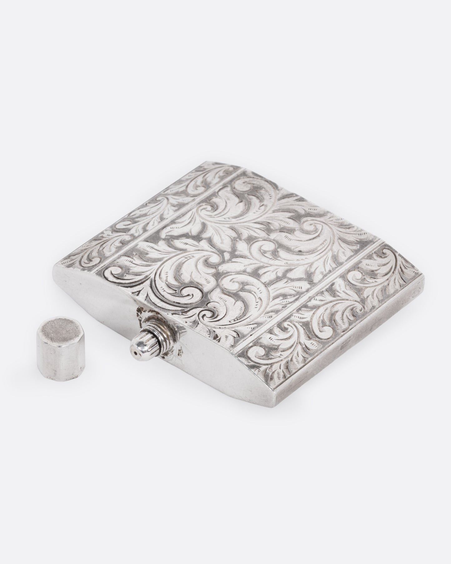A silver perfume bottle with winding botanical etchings and a slim square silhouette, shown from the side with its cap next to it.