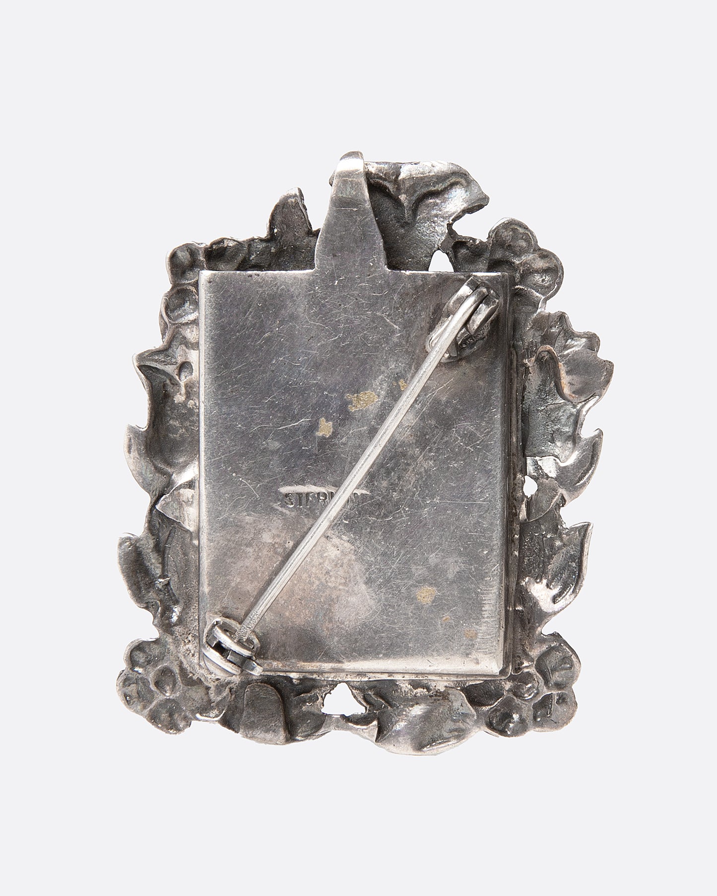 An ornate  silver picture frame brooch with a botanical motif around a rectangular opening, shown from the back.